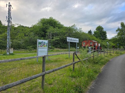 Landschaft mit Weg, Zaun, Mobilfunkmast und Schildern in grüner Umgebung.